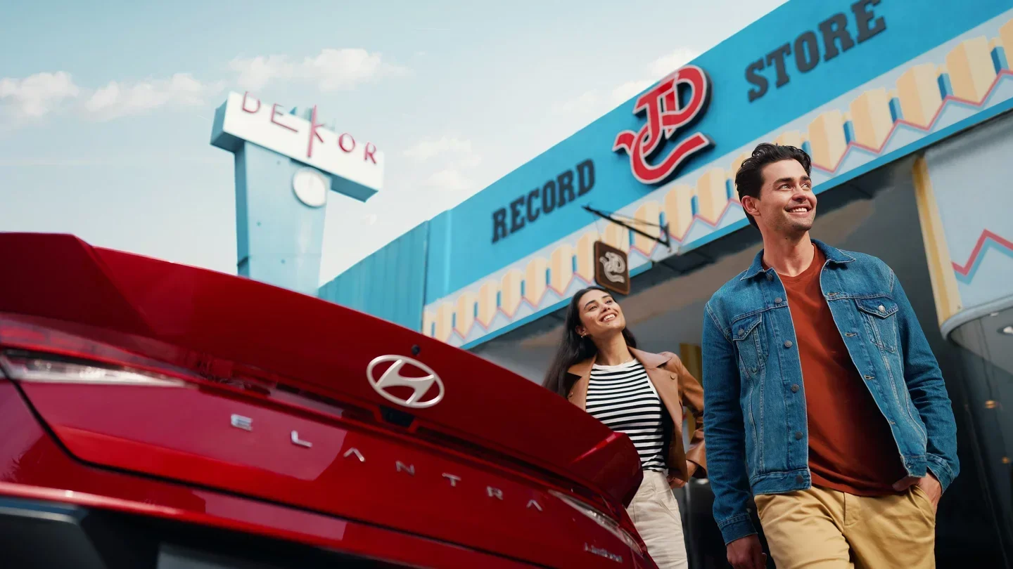 A couple walking past a red Hyundai Elantra outside a retro record store, the kind of everyday-life setting the Elantra fits into