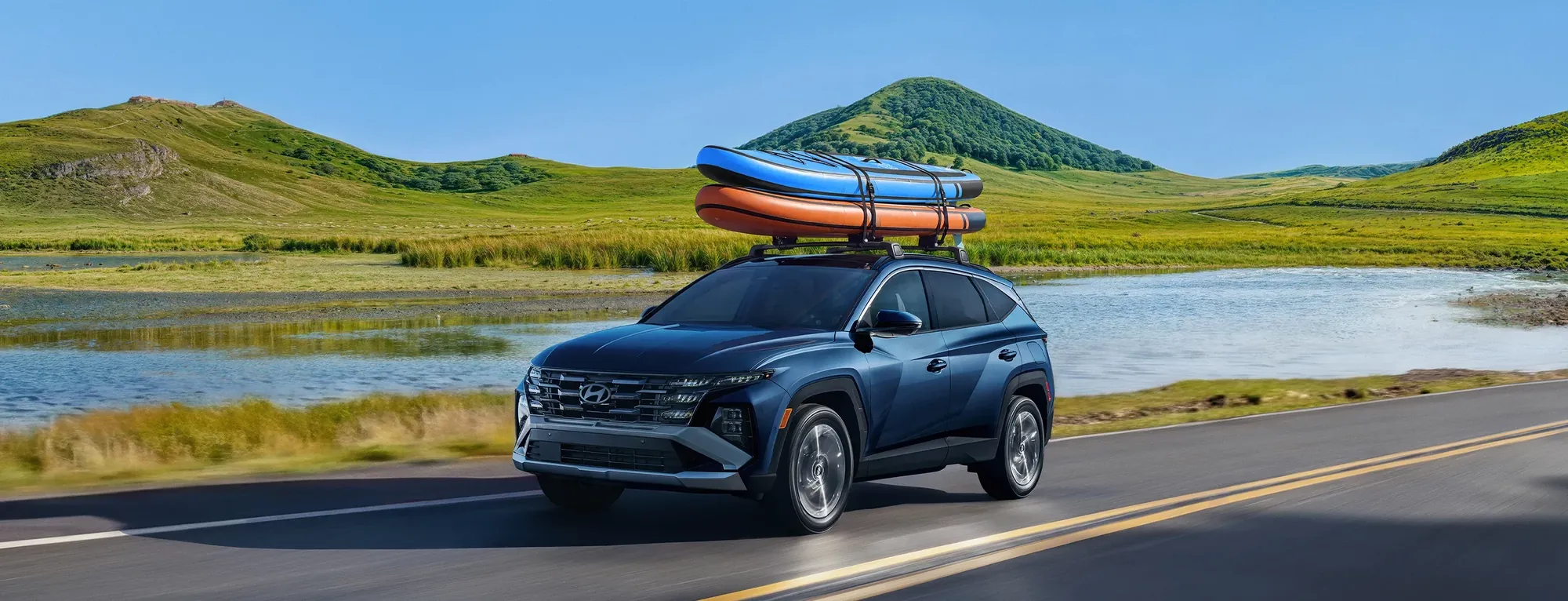 2026 Hyundai Tucson with kayaks on the roof rack driving past a lake, a compact SUV sold at Dalton Hyundai National City
