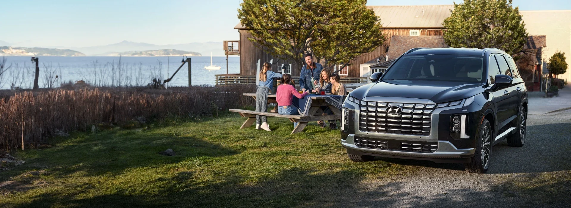 A Hyundai Palisade parked by a lake while a family picnics nearby.